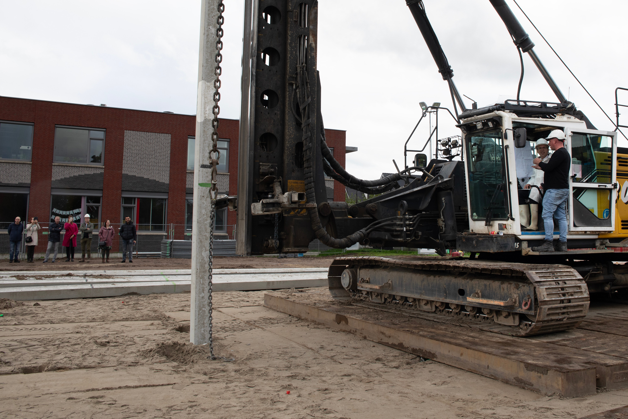 Wethouder zit in een kraan en slaat de eerste paal. Belangstellenden staan aan de zijkant te kijken.