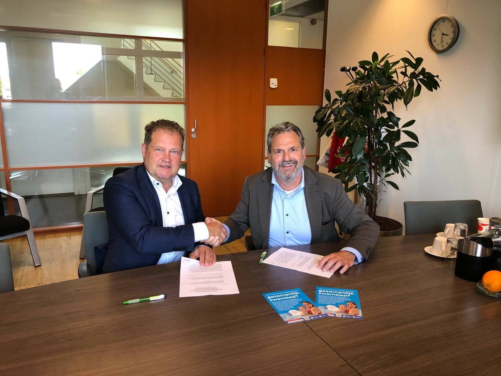 Wethouders zitten aan tafel en geven elkaar een hand. Getekende Documenten liggen op tafel.